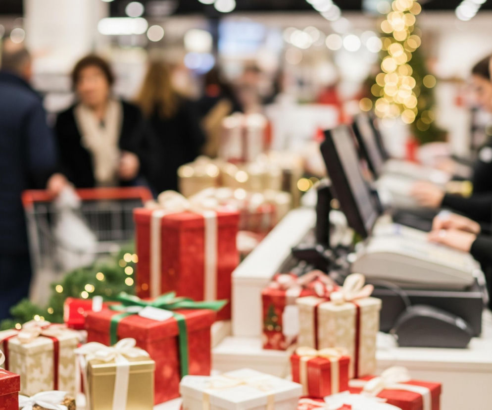 Na pierwszym planie, w dolnej części obrazu, znajduje się ekspozycja z licznymi prezentami zapakowanymi w czerwony, złoty i biały papier, przewiązanymi satynowymi wstążkami. W tle, po prawej stronie, widać trzy kobiety w ciemnych strojach, obsługujące kasy fiskalne. Po lewej stronie i w centrum, w głębi sklepu, widać rozmyte sylwetki innych klientów oraz regały z produktami i ozdobioną lampkami choinkę. Oświetlenie w tle tworzy liczne, rozmyte, jasne punkty świetlne.