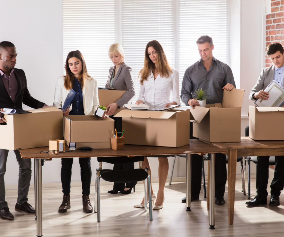 Grupa pracowników pakujących swoje rzeczy do kartonów, symbolizująca masowe zwolnienia w branży tech. Smutne twarze osób opuszczających miejsca pracy po redukcji etatów. Więcej o wpływie AI na rynek pracy przeczytasz na Super Biznes.