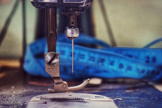 Stopka i igła maszyny do szycia z nitką, na blacie fragment metalowego blatu maszyny, w tle niebieska taśma miernicza. Zdjęcie symbolizuje usługi krawieckie, które Żabka wprowadza w swojej ofercie, o czym przeczytasz na portalu Super Biznes.