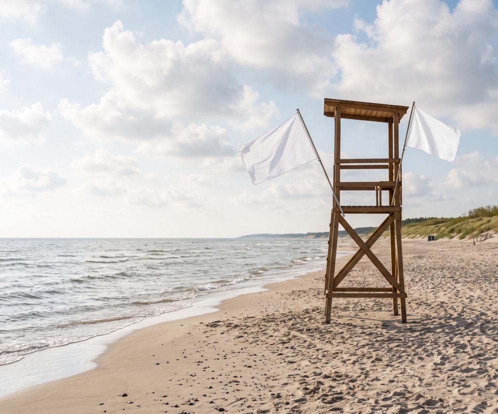 Drewniana wieża ratownika z dwiema białymi flagami na pustej plaży. O bezpiecznej kąpieli w Łazach czytaj w SE Szczecin.