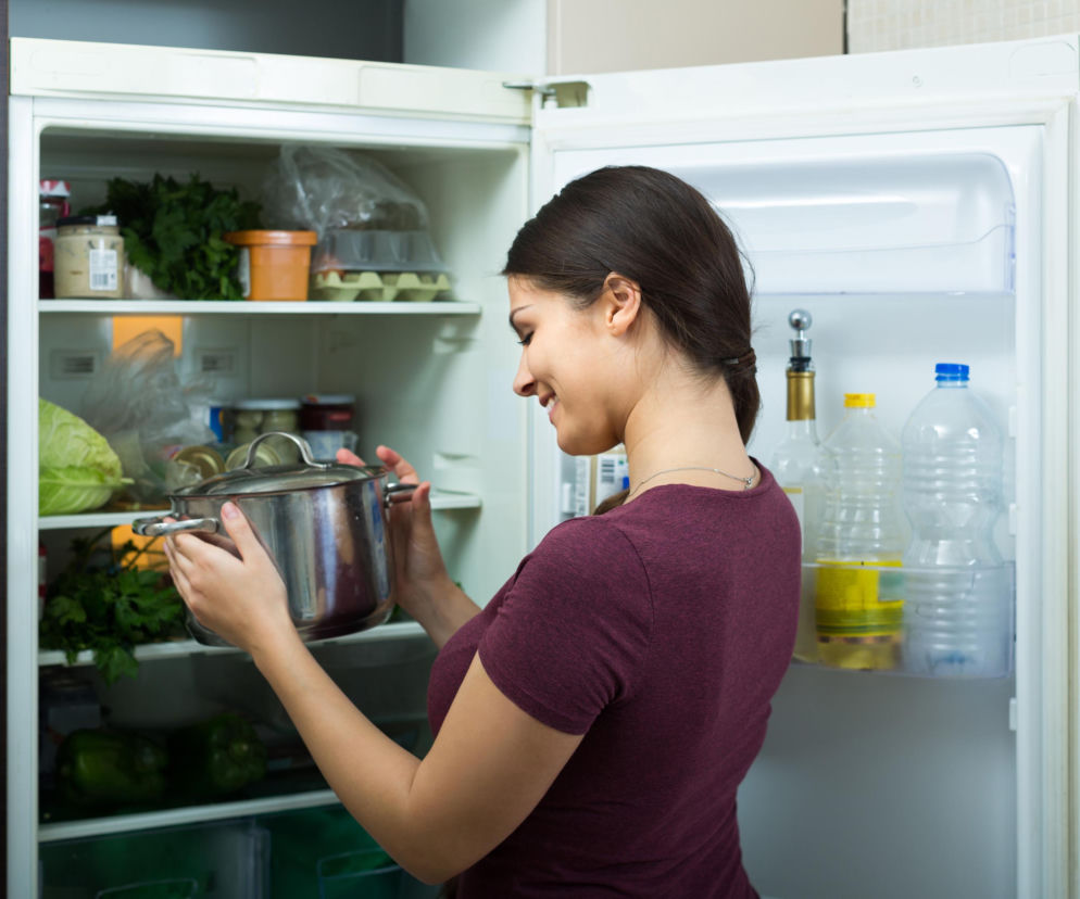 Młoda, uśmiechnięta kobieta w bordowej koszulce wkłada gorący garnek z parującym jedzeniem do otwartej, pełnej żywności lodówki, co jest zgodne z nowymi wytycznymi CDC dotyczącymi przechowywania posiłków. O bezpiecznym chłodzeniu jedzenia przeczytasz na Poradnik Zdrowie.