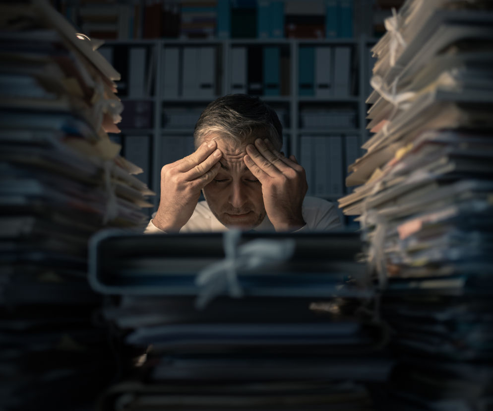 Mężczyzna z dłońmi na skroniach, wyraźnie zmęczony lub zestresowany, siedzi przy biurku otoczony stosami dokumentów, co symbolizuje wyczerpującą pracę, taką jak nocne zmiany. Więcej o wpływie pracy zmianowej na zdrowie przeczytasz na portalu Poradnik Zdrowie.