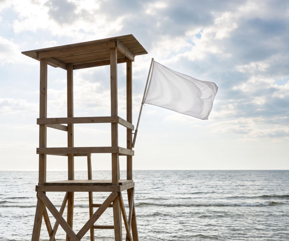 Drewniana wieża ratownicza na plaży z białą flagą na tle morza i nieba. O warunkach w Grzybowie pisze SE Szczecin.