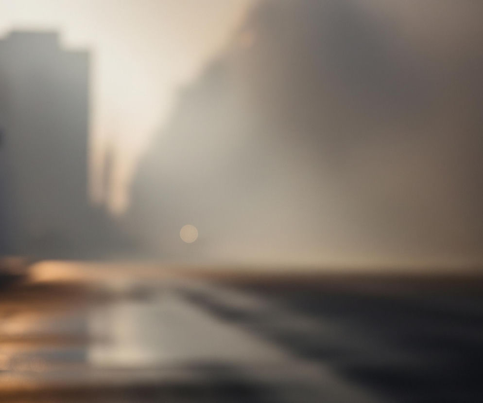 Rozmyty obraz przedstawia mglisty miejski krajobraz oświetlony ciepłym światłem. Po lewej stronie widać ciemną ścianę budynku, przechodzącą w niewyraźne, ciemne sylwetki innych konstrukcji, zanurzonych w jasnej mgle. Środek i prawa strona obrazu to szarobrązowa mgła, a na środku, bliżej dołu, wyróżnia się jasny, rozmyty punkt światła. Dolna część obrazu ukazuje mokrą, odbijającą światło powierzchnię, prawdopodobnie ulicę lub chodnik, z refleksami w odcieniach złota i brązu.