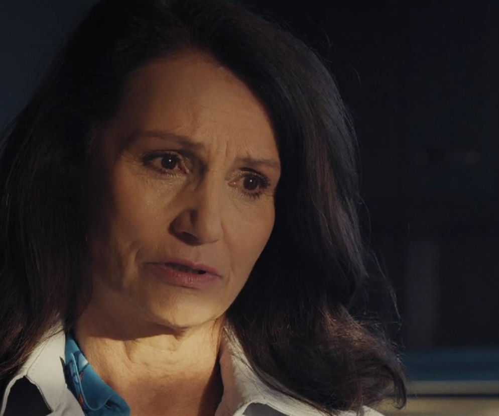 Małgorzata Pieczyńska jako Aleksandra w serialu M jak miłość w zbliżeniu na twarz, z zamyślonym, lekko zatroskanym wyrazem. Kobieta ma ciemne włosy, ubrana jest w białą koszulę z niebieskim akcentem. O jej wątpliwościach przeczytasz na Super Seriale.