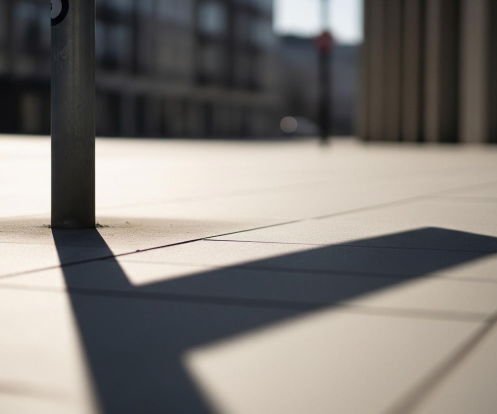 Na pierwszym planie, po lewej stronie kadru, wznosi się pionowy, metalowy słup w kolorze szarym. U jego podstawy widoczne są nierówności i ciemniejsza, granulowana powierzchnia styku z podłożem. Od słupa rozciąga się długi, ciemny cień w kształcie prostokąta, biegnący ukośnie w dół i w prawo przez jasną, kafelkową powierzchnię. Podłoże składa się z dużych, kwadratowych płytek w odcieniach beżu i jasnej szarości, z wyraźnymi liniami fug, oświetlonych ciepłym, bocznym światłem. W tle, rozmyte i niewyraźne, rysują się kontury budynków o ciemnych fasadach i oknach, zdominowane przez brązy i szarości, oraz pojedynczy, niewyraźny czerwony obiekt.