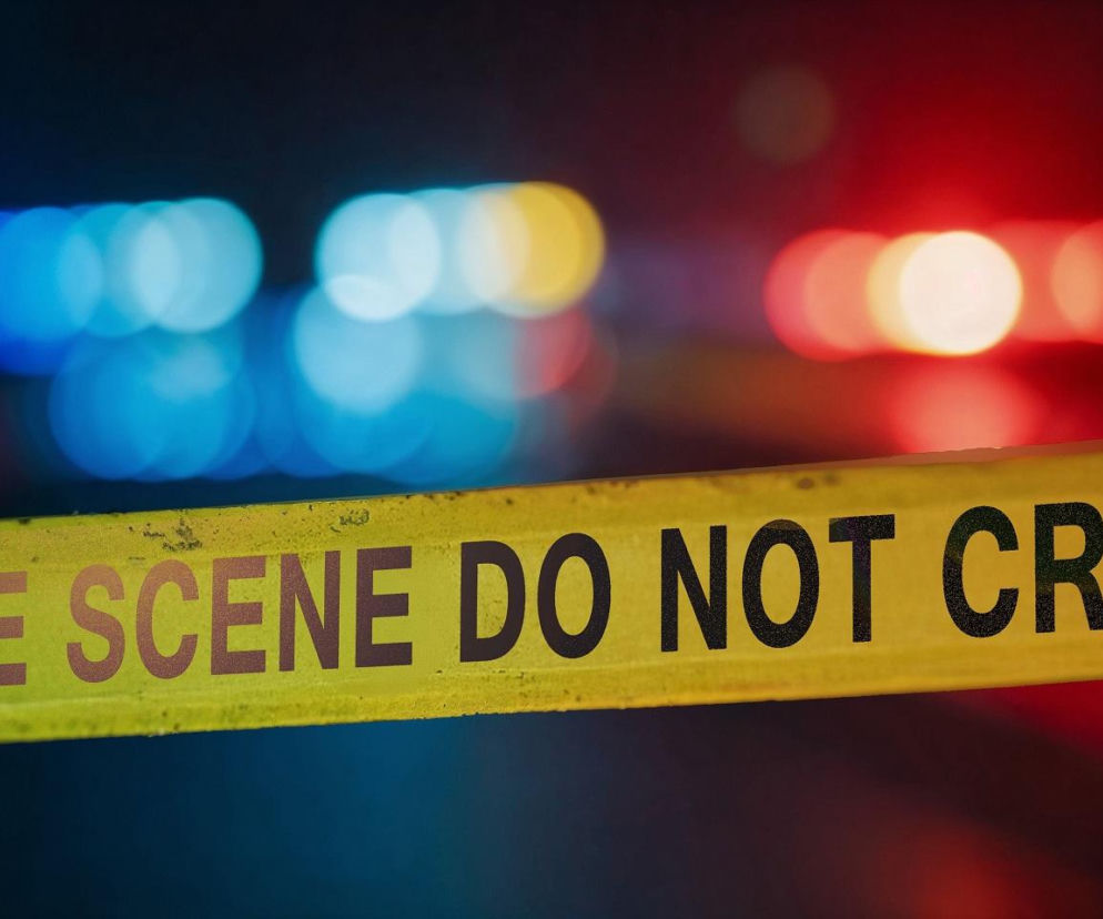 Żółta taśma policyjna z napisem „CRIME SCENE DO NOT CROSS” na pierwszym planie, rozmyte czerwone i niebieskie światła pojazdów ratunkowych w tle, symbolizujące tragiczne wydarzenie na koncercie, o którym możesz przeczytać na naszym portalu.