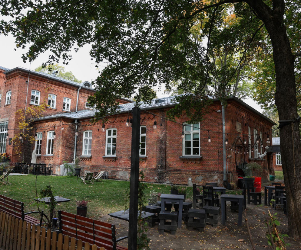 Zabytkowy budynek z czerwonej cegły otoczony drzewami i stolikami kawiarnianymi. O historii Szpitala Ujazdowskiego czytaj w SE.