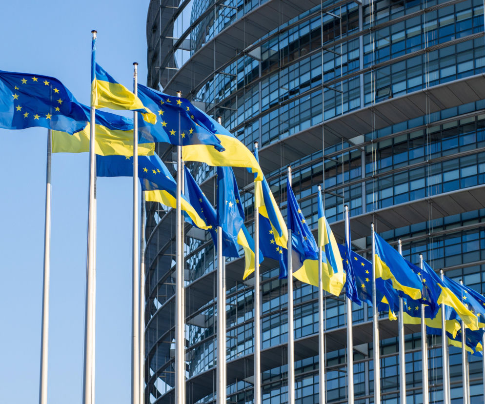 Flagi Unii Europejskiej i Ukrainy powiewające na wietrze przed nowoczesnym budynkiem. Symboliczne przedstawienie współpracy UE z Ukrainą w kontekście uruchomienia pomocy finansowej. Więcej na ten temat przeczytasz na portalu Super Biznes.