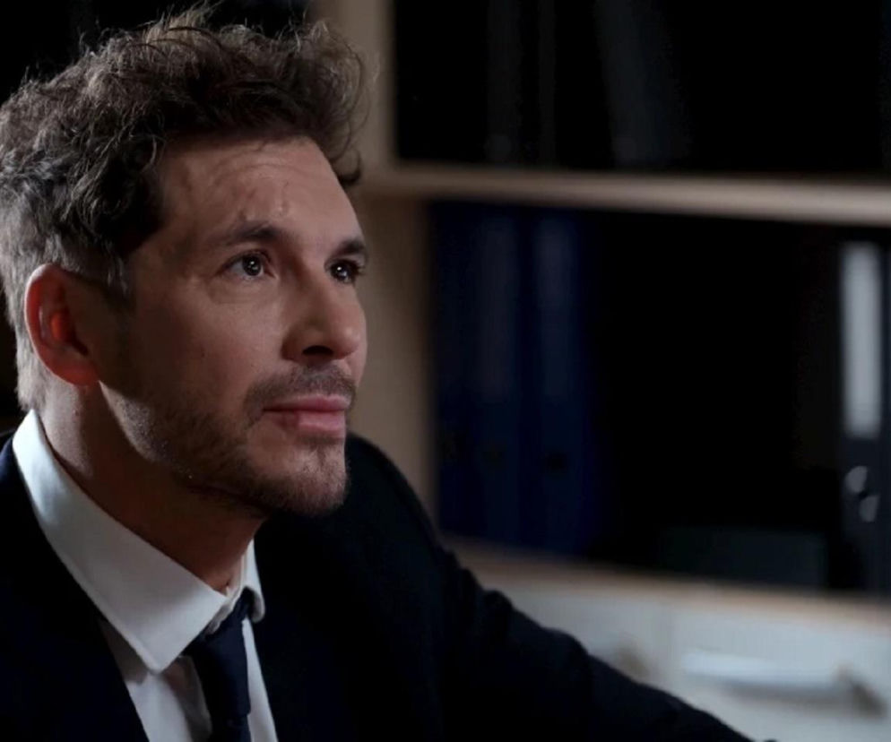 Mikołaj Roznerski jako Marcin Chodakowski w garniturze z krawatem, z zamyślonym wyrazem twarzy, spoglądający w prawą stronę. To kluczowa scena z serialu M jak miłość, gdzie bohater stawia czoła nowym wyzwaniom zawodowym, a o jego losach przeczytasz więcej na Super Seriale.