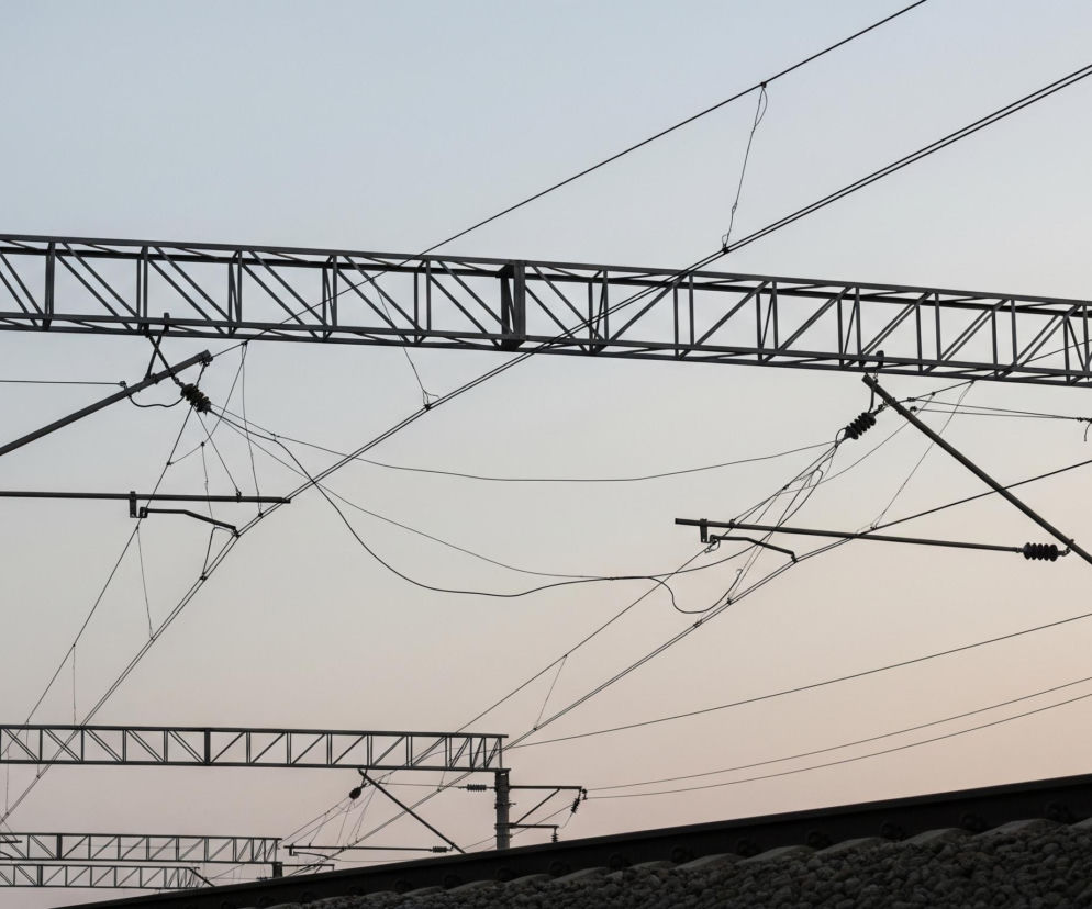 Ciemne, metalowe konstrukcje wsporcze i liczne przewody trakcji kolejowej na tle jasnego, zachodzącego nieba. Dominują tu szarości i czernie sylwetek słupów, wsporników kratownicowych oraz gęstej sieci przewodów elektrycznych, które krzyżują się i rozciągają na różnej wysokości. W dolnej części widoczne są fragmenty ciemnych torów kolejowych ułożonych na jasnym tłuczniu. Całość skąpana jest w delikatnym, rozbielonym świetle zgaszonego różu i bladego błękitu, tworząc atmosferę zmierzchu.