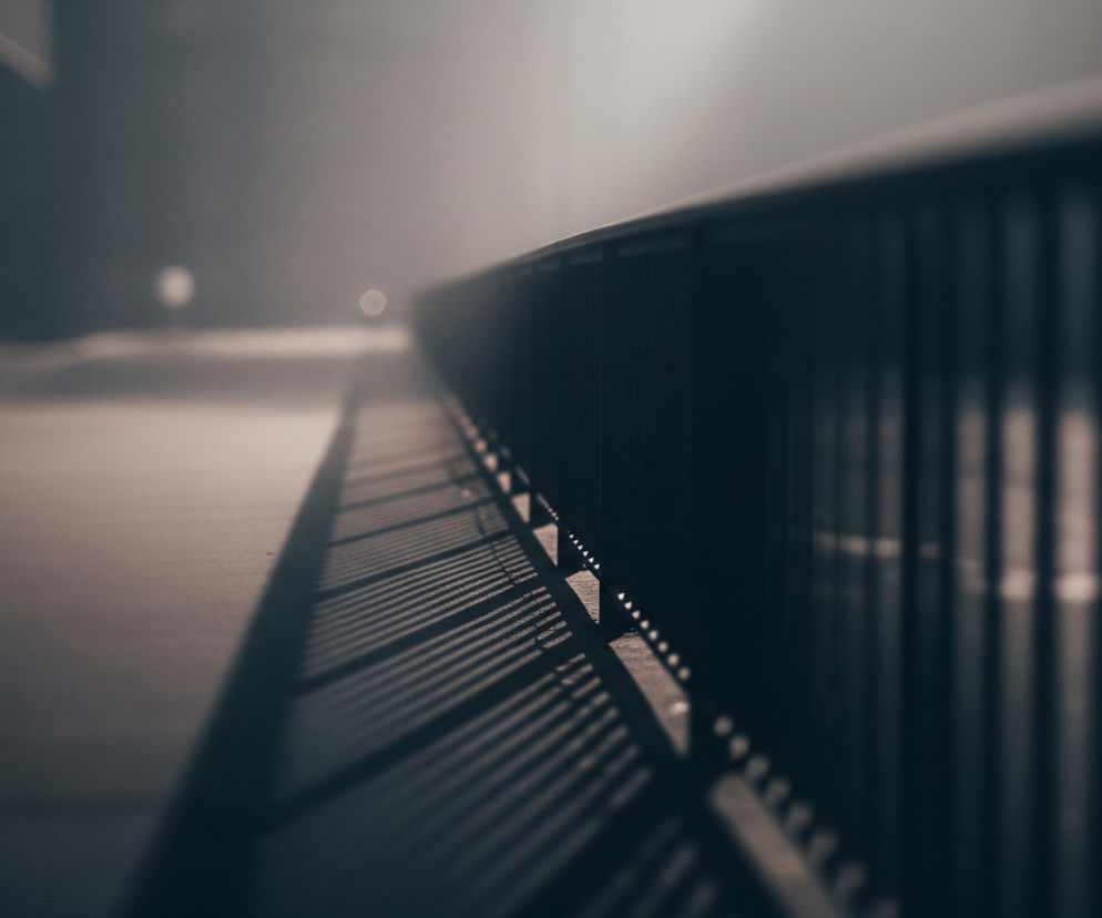 Metalowa barierka biegnie ukośnie w górę, zajmując prawą część kadru, z wyraźnym rozmyciem tła. Rzucane przez nią cienie tworzą równoległe, ciemne pasy na jasnym chodniku po lewej stronie. W tle, w oddali, widać rozmyte, jasne punkty światła na szarym, zamglonym tle, a także zarys ciemnych, pionowych słupów. Kolorystyka zdjęcia jest zdominowana przez ciemne, stonowane szarości i brązy.