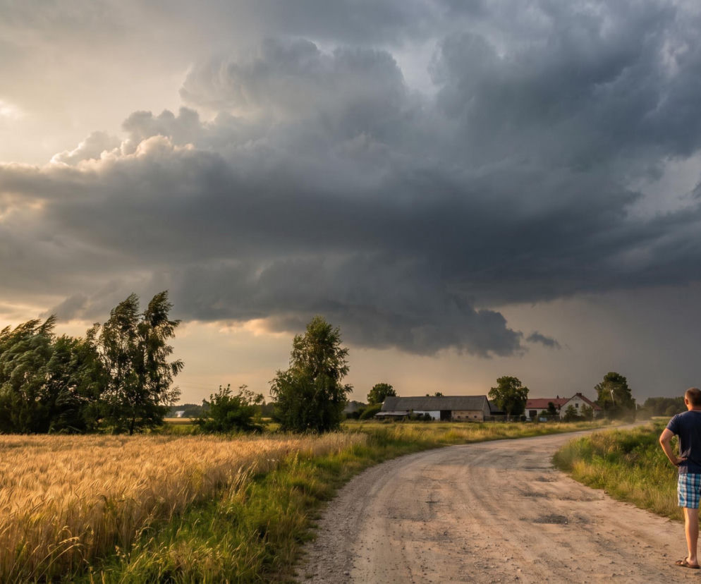 Dwie osoby stoją na wiejskiej drodze gruntowej, obserwując zbliżającą się, groźną nawałnicę z ciemnymi chmurami burzowymi nad polami zbóż i zabudowaniami, co symbolizuje ekstremalne zjawiska pogodowe w Polsce. Więcej informacji o burzach i upałach znajdziesz na naszym portalu.
