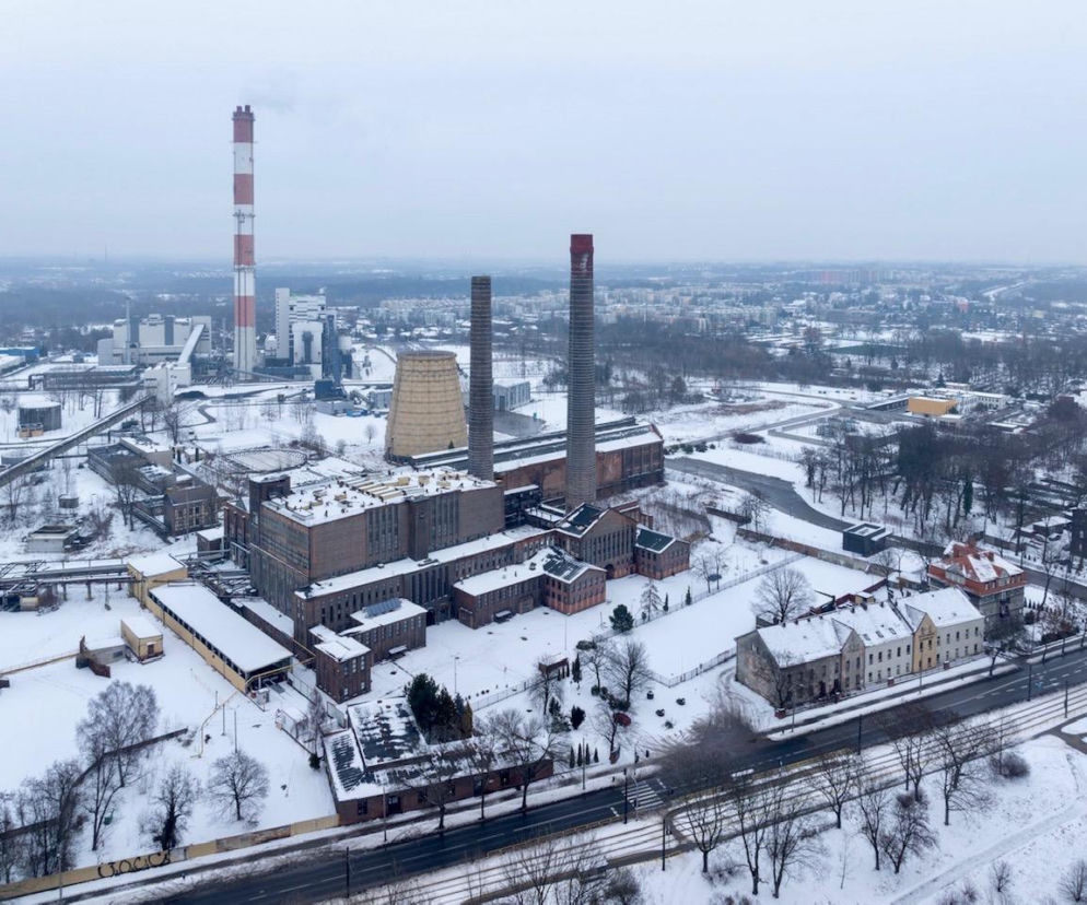 Zimowy krajobraz Elektrociepłowni Zabrze Grupy Arche, obejmujący zabytkowe budynki i kominy pod śniegiem. Na pierwszym planie zabudowania otoczone ośnieżoną roślinnością i droga, w tle miejska panorama. O rewitalizacji obiektu przeczytasz na Architektura Murator Plus.