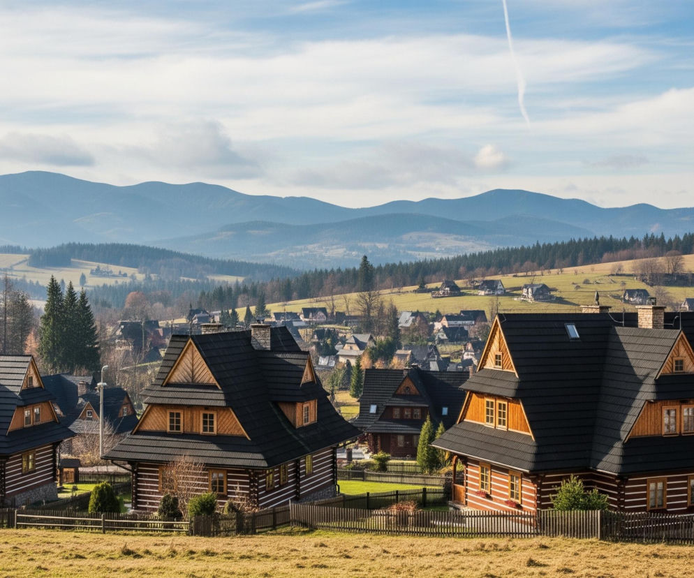 Pierwszy plan wypełnia brązowo-żółta trawa, za którą widać drewniane płoty. Po lewej stronie znajdują się trzy drewniane domy w tradycyjnym stylu, z ciemnymi dachami i ozdobnymi, jasnobrązowymi elementami na szczytach. Za nimi rozpościera się widok na rozległą dolinę z licznymi, podobnymi domami, otoczonymi zielonymi i brązowymi polami oraz pasmami ciemnozielonych drzew iglastych. W tle wznoszą się rzędy zalesionych gór o niebieskawych odcieniach, nad którymi rozciąga się częściowo zachmurzone niebo z widocznym białym smugą kondensacyjną.