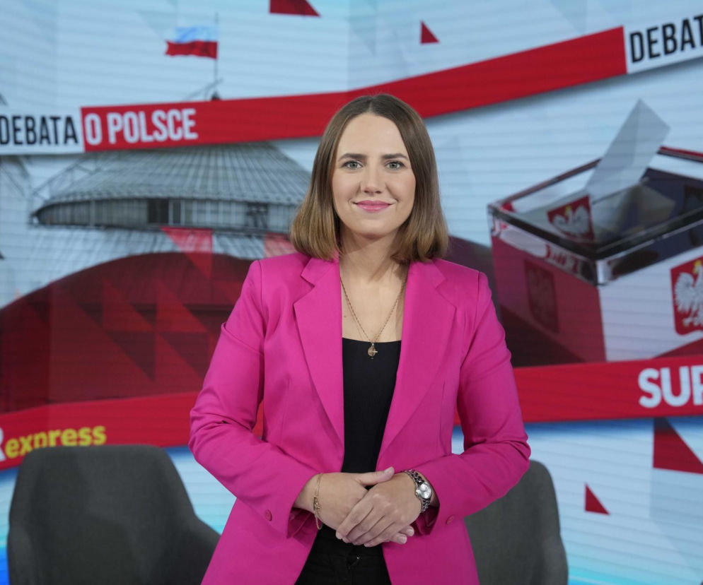 Europosłanka Konfederacji Anna Bryłka w różowej marynarce i czarnym topie, z rękami splecionymi na wysokości brzucha, stoi przed ścianką z napisem DEBATA O POLSCE i motywami związanymi z wyborami. O jej oburzeniu na patologię w Szpitalu Południowym przeczytasz na naszym portalu.