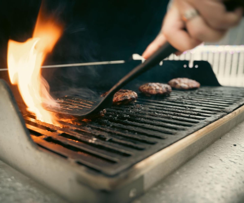 Ręka osoby obracającej łopatką burgery na ruszcie, z którego buchają płomienie. O czyszczeniu grilla czytaj na SE.