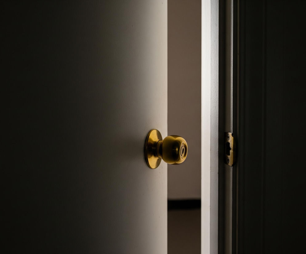 Drzwi, częściowo otwarte, dominują w kadrze, ukazując jasną, kremową barwę na zewnętrznej powierzchni i ciemniejszy odcień na wewnętrznej krawędzi. Na centralnej osi drzwi znajduje się okrągła, mosiężna klamka z otworem na klucz w środku, odbijająca światło. Po prawej stronie widać futrynę drzwi, również w jasnym kolorze, z widocznym, prostokątnym mechanizmem zamka, również w odcieniu mosiądzu, z ciemnymi otworami na śruby. Tło za drzwiami pozostaje w głębokim cieniu, jedynie delikatne światło przenika przez szczelinę między drzwiami a futryną, tworząc pionowy pas jaśniejszej barwy po prawej stronie.