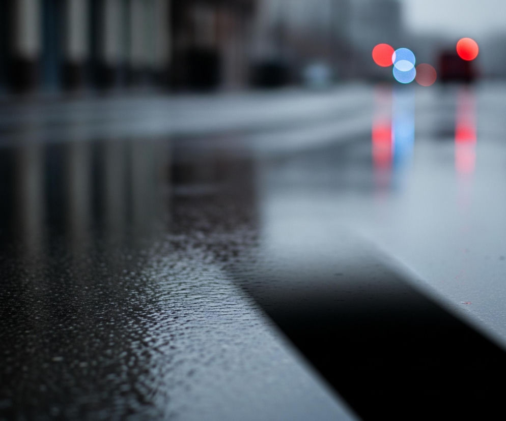 Mokra, ciemnoszara nawierzchnia drogi z widocznymi refleksami światła zajmuje większą część kadru. W dolnej części obrazu widoczny jest szeroki, czarny pas, prawdopodobnie poziome oznakowanie drogowe, biegnący ukośnie od lewej do prawej. Na drugim planie, rozmyte, widoczne są intensywnie czerwone i niebieskie światła, prawdopodobnie pochodzące z sygnalizacji świetlnej lub pojazdów uprzywilejowanych, które odbijają się w kałużach. Tło jest niewyraźne, zarysowują się jedynie ciemne, pionowe kształty budynków.