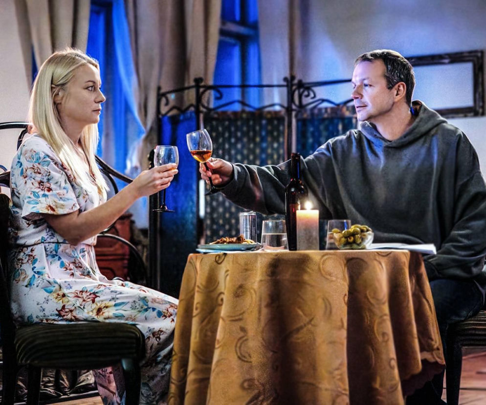 Mateusz i Emilka z serialu Pierwsza miłość siedzą przy stole, wznosząc toast winem. Emilka ma na sobie kwiecistą sukienkę i wyraźnie widać niebieskie kajdanki na jej dłoni. W tle ciemne okno i stylizowane meble. Więcej o ich losach przeczytasz na Super Seriale.