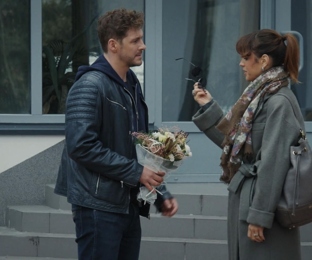 Mikołaj Roznerski jako Marcin z bukietem kwiatów i Michalina Sosna jako Kama z okularami przeciwsłonecznymi w ręku, oboje w szarych płaszczach, na tle szarego budynku ze szklanymi drzwiami i schodami, podczas konfrontacji. Więcej informacji o serialu znajdziesz na Super Seriale.