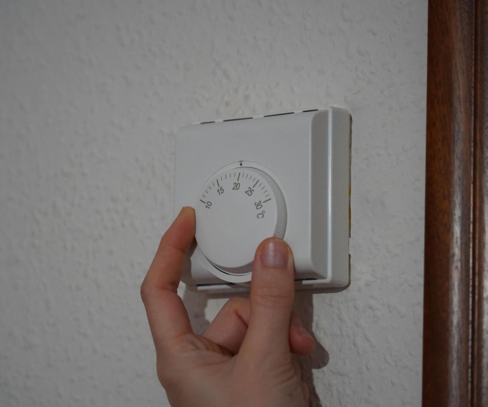 Kobieca dłoń reguluje biały termostat ścienny, ustawiając temperaturę. To wizualizacja problemu zbyt ciepłej sypialni, która zagraża sercu, o czym można przeczytać na portalu Poradnik Zdrowie.