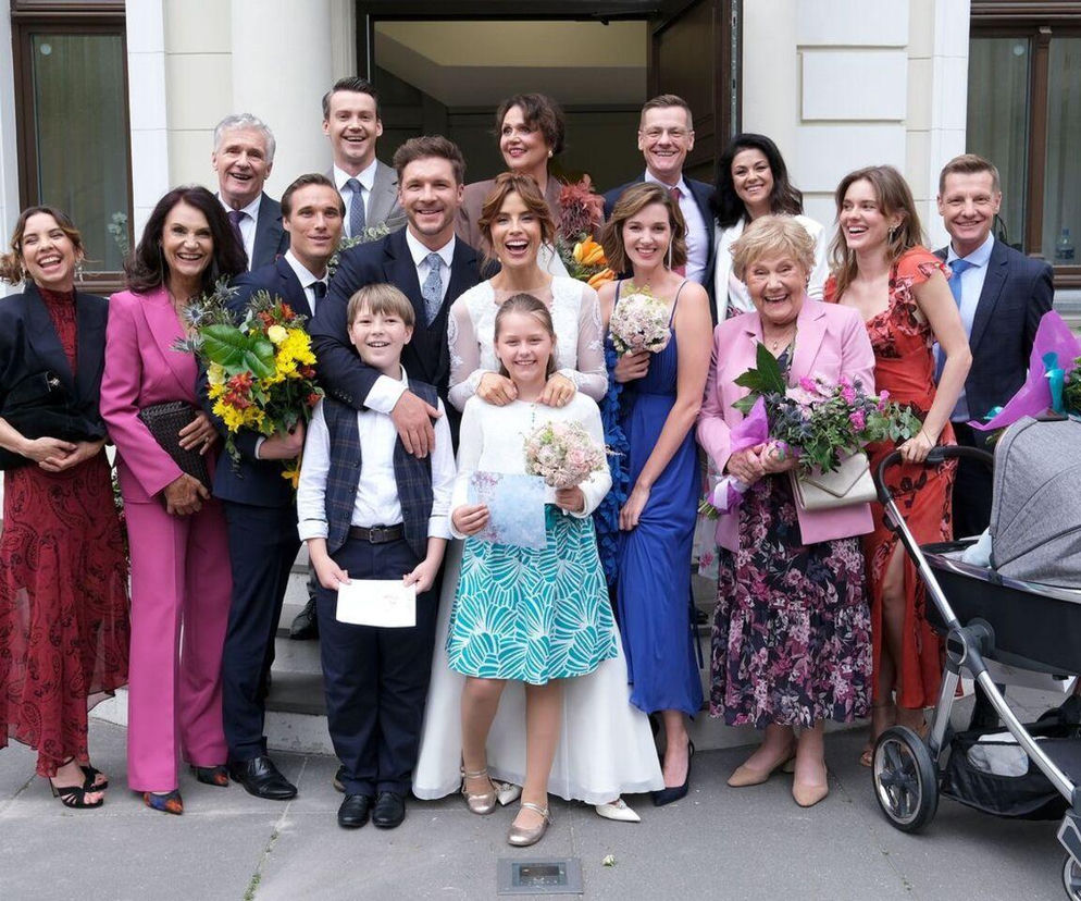 Duża grupa gości weselnych ze ślubu Kamy i Marcina z serialu M jak miłość pozuje do zdjęcia przed urzędem stanu cywilnego. Na pierwszym planie młoda para z dziećmi, uśmiechnięta Barbara Mostowiak oraz inni aktorzy serialu M jak miłość. Więcej o tym odcinku przeczytasz na stronie Super Seriale.