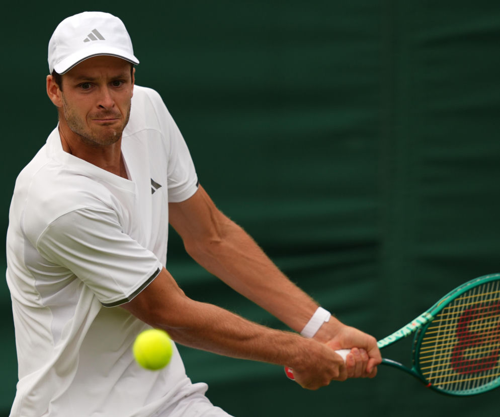 Hubert Hurkacz w białym stroju sportowym i czapce z daszkiem, z determinacją odbija piłkę tenisową na tle zielonego kortu podczas meczu Wimbledonu. O jego zwycięstwie nad Casperem Ruudem przeczytasz więcej na naszym portalu.
