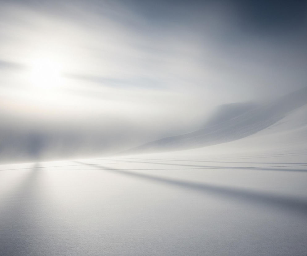 Zasłana śniegiem dolina rozciąga się w kierunku horyzontu pod zamglonym niebem. Po lewej stronie górny róg jest rozświetlony przez jasne słońce, które przenika przez białą mgłę, rzucając wydłużone cienie na płaski, śnieżny krajobraz. Po prawej stronie wznosi się góra, częściowo ukryta we mgle, z jej zboczami pokrytymi śniegiem, przechodzącymi w ciemniejsze odcienie szarości w górnej części kadru.