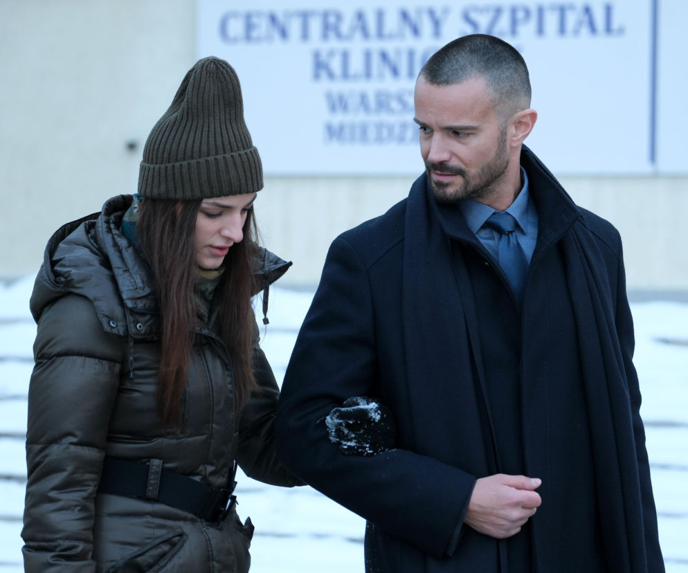 Kasia (Paulina Lasota) w oliwkowej czapce i zimowej kurtce, oparta o ramię Sowińskiego (Mikołaj Krawczyk) w ciemnym płaszczu i krawacie, przed wejściem do kliniki. Zdjęcie ilustruje sceny z odcinka 1935 M jak miłość. Więcej o serialu przeczytasz na Super Seriale.