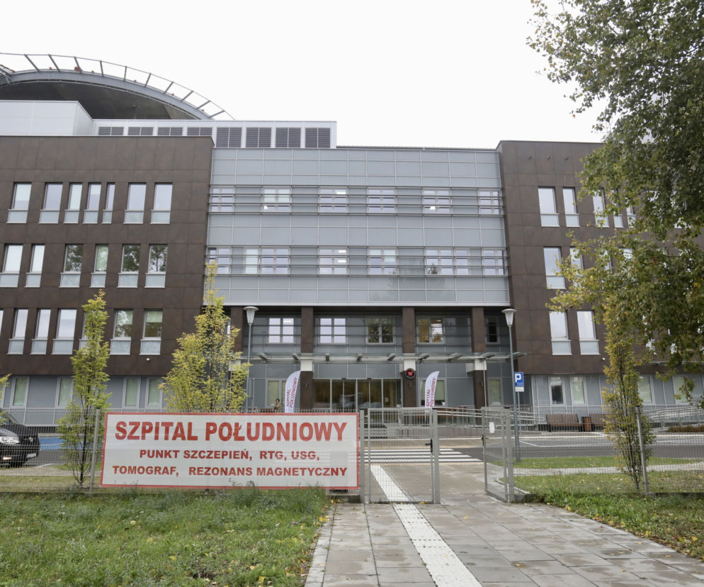 Szpital Południowy w Warszawie, nowoczesny budynek z ciemną elewacją i szklanymi fasadami, z lądowiskiem dla helikopterów na dachu. Przed wejściem widoczny baner informacyjny SZPITAL POŁUDNIOWY, podkreślając placówkę, o której czytasz na naszym portalu.