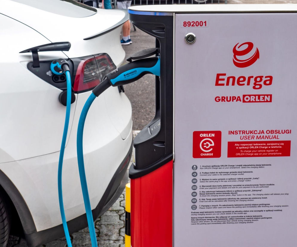 Biały samochód elektryczny Tesla ładowany na stacji Energa Grupa ORLEN, z widocznym panelem sterowania ładowarki. Kierowcy elektryków mogą liczyć na obniżki cen ładowania, o czym pisze Super Biznes.
