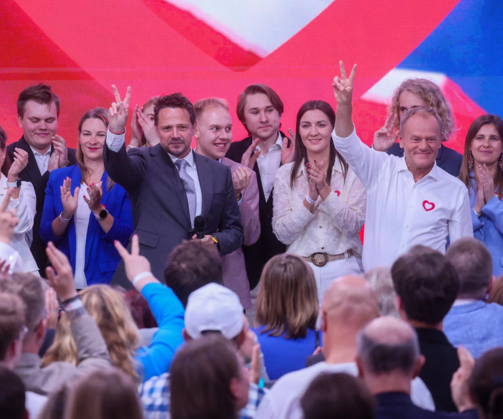 Donald Tusk z uśmiechem i uniesioną dłonią w geście zwycięstwa na tle publiczności, otoczony politykami Koalicji Obywatelskiej, m.in. Rafał Trzaskowski również pokazujący ten gest. Wszyscy wydają się zadowoleni. Więcej o sondażach przeczytasz na naszym portalu.