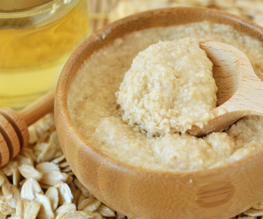 Gęsta owsiana pasta w bambusowej misce, obok słoik z miodem i drewniana łyżka do miodu, na rozsypanych płatkach owsianych. To domowy sposób na kojenie podrażnionej skóry, o którym przeczytasz więcej na Poradnik Zdrowie.