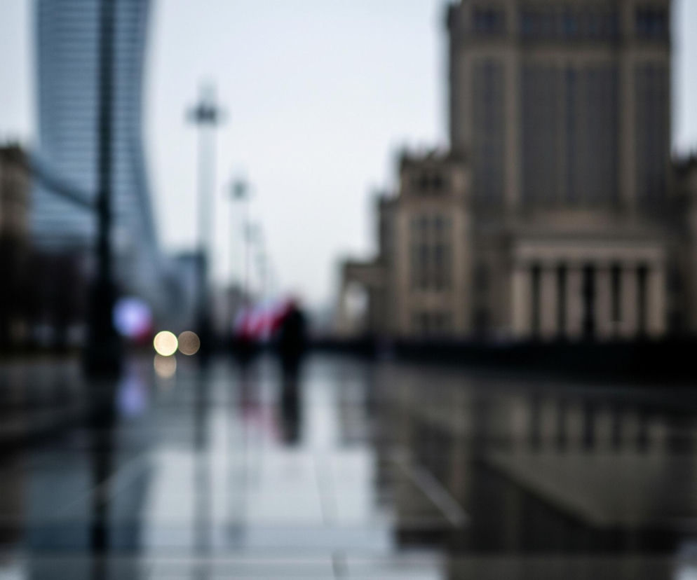 Rozmazany, miejski krajobraz, dominują szare i brązowe odcienie. Na pierwszym planie widoczna jest mokra powierzchnia, prawdopodobnie ulica lub chodnik, odbijająca światła i zarysy budynków. W oddali, po prawej stronie, wznosi się duży, ciemny budynek z kolumnami, a po lewej fragment innego, nowoczesnego, szklanego wieżowca. Pomiędzy budynkami widać rozmyte sylwetki ludzi, w tym jedna postać pod czerwonym parasolem, oraz jasne, okrągłe punkty światła, odzwierciedlające się w mokrej nawierzchni.