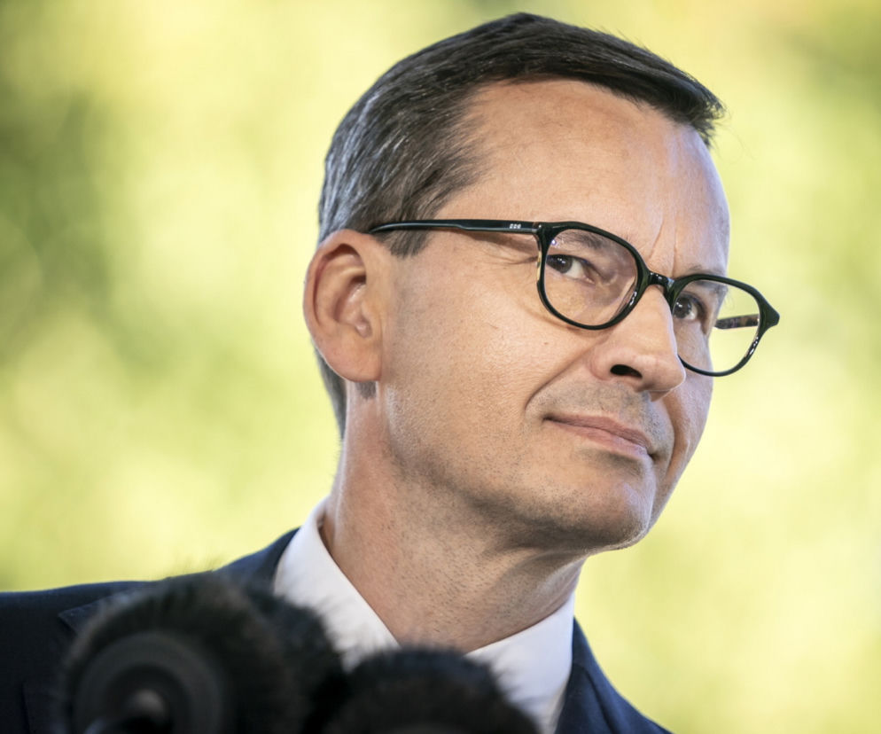 Mateusz Morawiecki w okularach, w garniturze i białej koszuli, patrzący w bok podczas wystąpienia. Zbliżenie na jego twarz, w tle rozmyta zieleń. Były premier prezentuje program Polska Jednej Prędkości, o którym więcej przeczytasz na Super Biznes.
