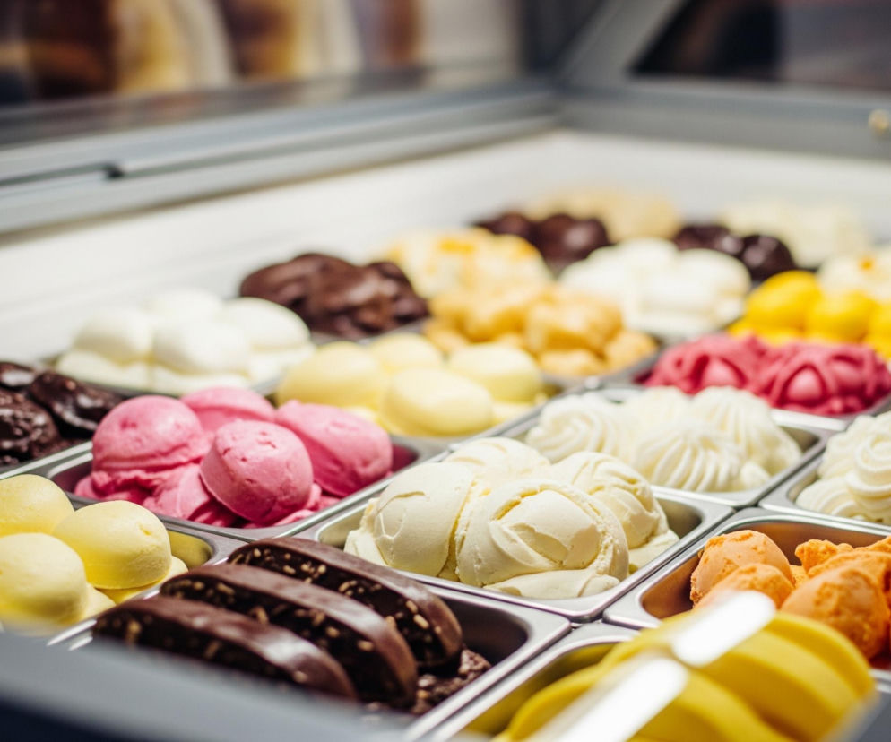 W centralnej części obrazu znajduje się witryna chłodnicza wypełniona metalowymi pojemnikami z różnokolorowymi lodami i deserami. W pierwszym planie po lewej stronie widoczne są gałki jasnożółtych lodów, a obok nich ciemnobrązowe batony pokryte czekoladą. Dalej w głąb witryny, po prawej stronie, ułożone są białe, udekorowane kremem w kształcie rozetek porcje deserów oraz pomarańczowe gałki lodów.

W dalszych rzędach pojemników widać różowe, ciemnoczerwone, jasnożółte, brązowe oraz kremowe lody i desery, niektóre z nich są gładkie, inne mają wyraźną teksturę. Tło jest rozmyte, ukazując jedynie górną część witryny i odbijające się w niej światło. Całość jest jasna i skupia się na bogactwie smaków i kolorów prezentowanych deserów.