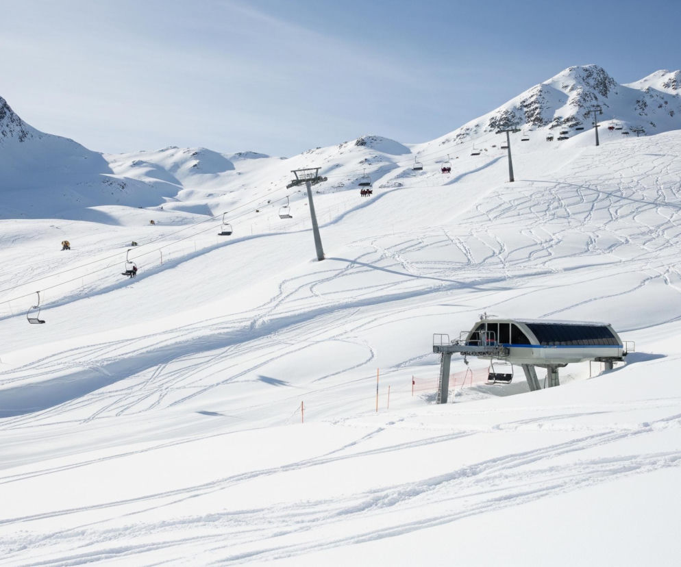 Ujęcie panoramiczne zimowego krajobrazu górskiego, dominują białe, ośnieżone stoki i szczyty, rozciągające się pod czystym, błękitnym niebem. Na pierwszym planie, w prawym dolnym rogu, widoczna jest stacja wyciągu narciarskiego z zadaszonym budynkiem, z którego odjeżdżają puste, czarne krzesełka, zawieszone na stalowej linie. Wzdłuż zbocza biegnie seria słupów podtrzymujących wyciąg krzesełkowy, a na niektórych krzesełkach widać siedzące sylwetki narciarzy, ubranych w ciemne kurtki. Na całym stoku, usianym licznymi śladami nart i desek snowboardowych, wyróżniają się pojedynczy narciarze zjeżdżający w dół oraz dwie armatki śnieżne w oddali, z których jedna, jasnobrązowa, znajduje się po prawej stronie.
