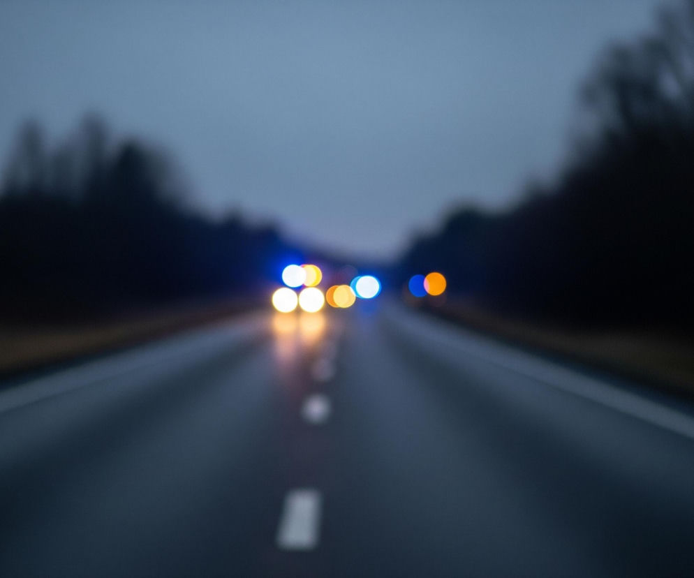 Asfaltowa droga rozciąga się w głąb obrazu, zajmując jego dolną połowę. Na jej środku widoczne są białe, przerywane linie, stające się coraz mniejsze i bardziej rozmyte w oddali. Na ciemnym asfalcie odbija się światło, co sugeruje mokrą powierzchnię. W tle, na horyzoncie, znajdują się rozmyte kształty drzew lub krzewów po obu stronach drogi, tworzące ciemną sylwetkę pod szarym, zachmurzonym niebem. Daleko na drodze widać rozmyte, kolorowe światła, w tym białe, pomarańczowe i niebieskie, wyglądające jak światła pojazdów lub sygnały świetlne.