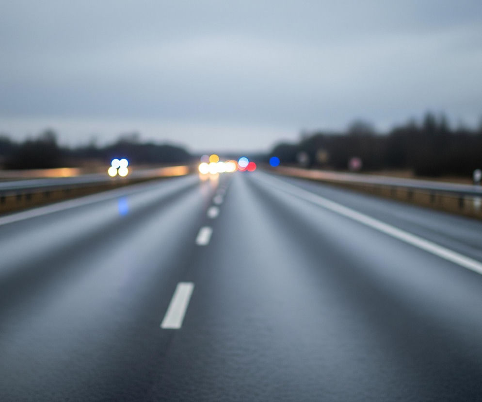 Szosa o wielu pasach biegnie przez środek kadru, widoczna od pierwszego planu po horyzont. Jej ciemna, mokra nawierzchnia odbija światła pojazdów i pochmurnego nieba, które zajmuje górną część obrazu. Białe, przerywane linie wyznaczają pasy ruchu w centrum, a ciągłe linie boczne oddzielają je od barierek ochronnych. Z lewej i prawej strony drogi rozciąga się niewyraźny krajobraz z ciemnymi drzewami i drogowskazami, z daleka widać rozmyte, kolorowe światła samochodów.