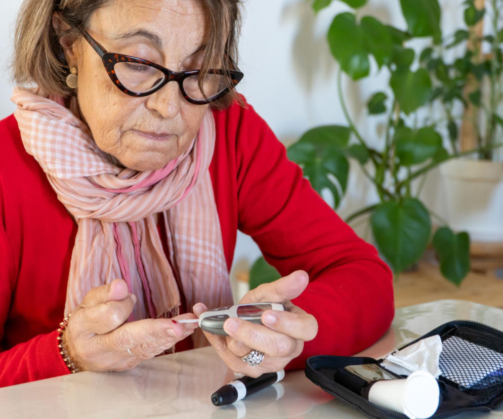 Starsza kobieta w okularach i czerwonym swetrze, z szalikiem na szyi, mierzy poziom cukru we krwi glukometrem, obok otwarte etui z akcesoriami medycznymi. Zdjęcie ilustruje temat zasiłku pielęgnacyjnego dla chorych na cukrzycę, o którym można przeczytać na Poradnik Zdrowie.