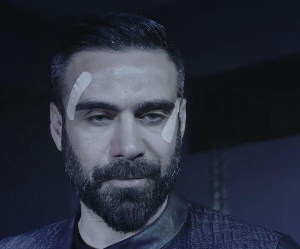 Mężczyzna z brodą i plastrami na skroniach oraz pod okiem, wyglądający na zmartwionego lub zmęczonego, w scenie z serialu Dziedzictwo, o którym więcej można przeczytać na Super Seriale. Obraz przedstawia ważny moment związany z Trucizną z 901. odcinka tureckiej telenoweli.