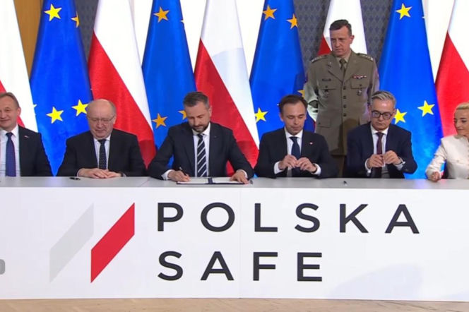 Grupa urzędników i polityków podczas podpisywania umów dotyczących programów obronnych, w tym Funduszy SAFE. Mężczyźni w garniturach, jedna kobieta w jasnym żakiecie. W tle flagi Polski i Unii Europejskiej. Informacje o takich wydarzeniach na Super Biznes.