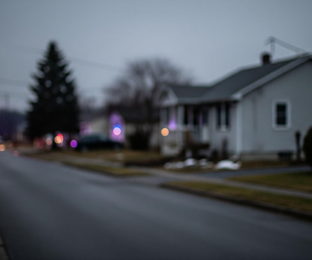 Rozmyta ulica, biegnąca od lewej strony ku górze obrazu, dominuje pierwszy plan. Na jej końcu widoczne są intensywnie świecące, rozmyte światła w kolorach czerwonym, niebieskim i pomarańczowym, sugerujące pojazd uprzywilejowany. Po prawej stronie drogi rozciąga się trawnik i chodnik, a za nimi szereg domów mieszkalnych o jasnych elewacjach i ciemnych dachach, otoczonych drzewami i krzewami. Ogólny nastrój jest stonowany i nieco ponury, z dominującymi odcieniami szarości i granatu.