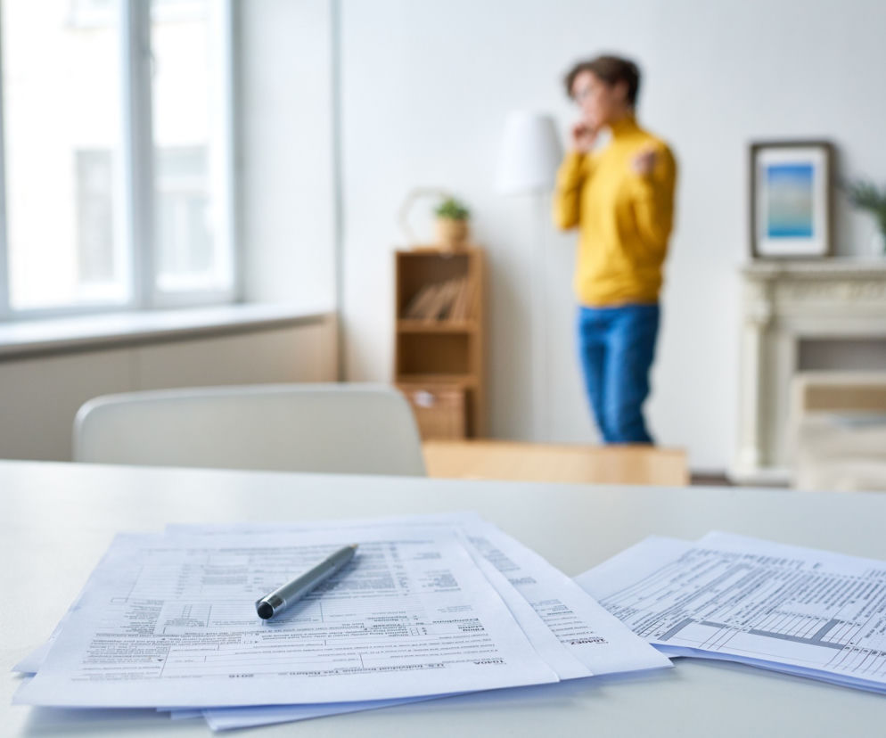 Dokumenty Małego ZUS Plus i długopis na biurku, w tle niewyraźna postać kobiety rozmawiającej przez telefon w domowym biurze. Zdjęcie symbolizuje nowe zasady i uproszczenia w Małym ZUS Plus, o których można przeczytać w portalu Super Biznes.