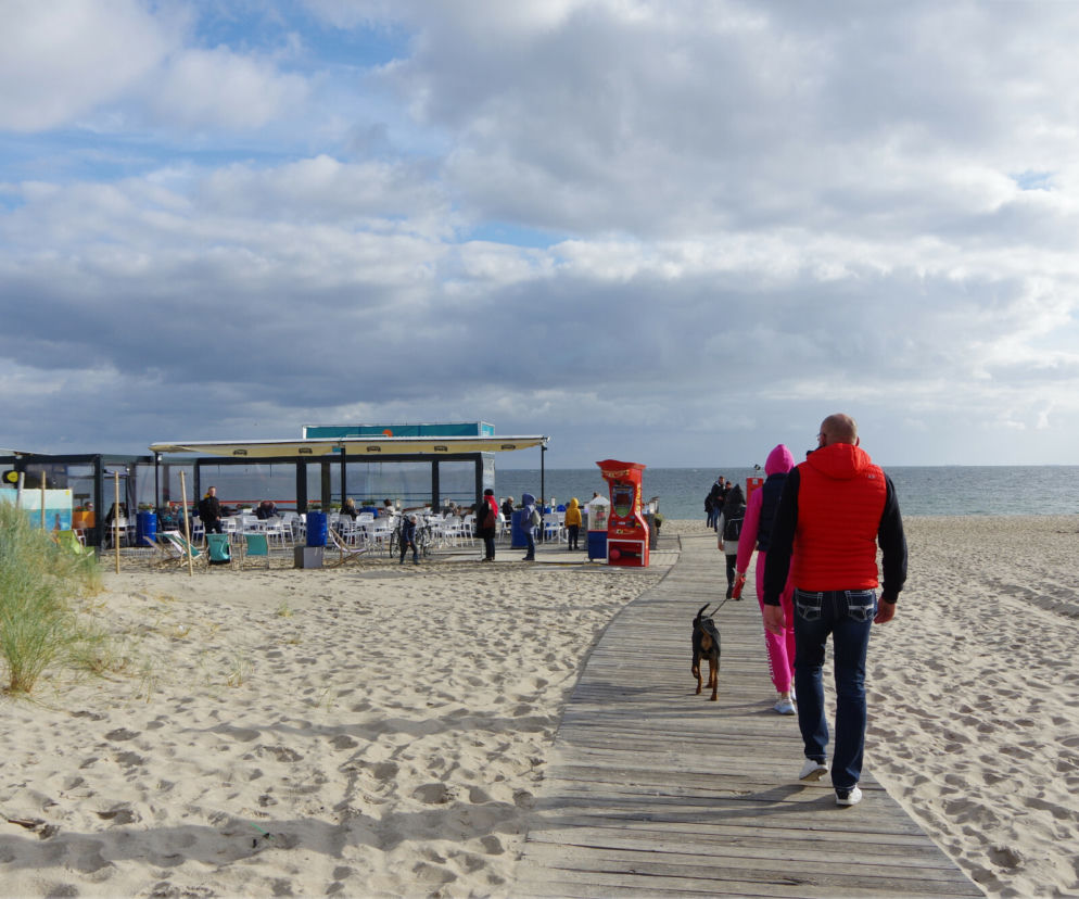 Osoby spacerujące drewnianą kładką po plaży jesienią. O niskich cenach wyjazdów nad morze przeczytasz na Eska.