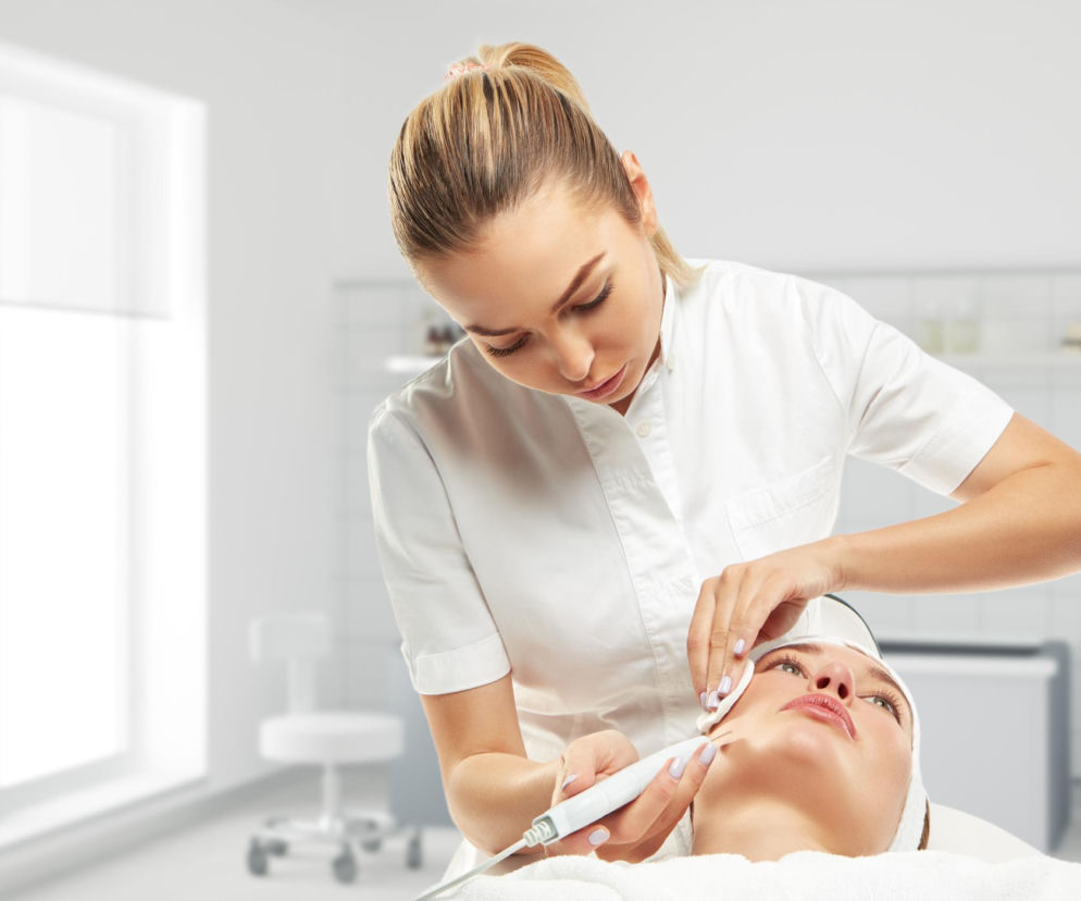 Kosmetyczka wykonuje zabieg na twarzy klientki, używając urządzenia do pielęgnacji skóry i wacika. Scena w gabinecie kosmetycznym przypomina o ważnych komunikatach Ministerstwa Zdrowia dotyczących medycyny estetycznej, o których można przeczytać na portalu Poradnik Zdrowie.