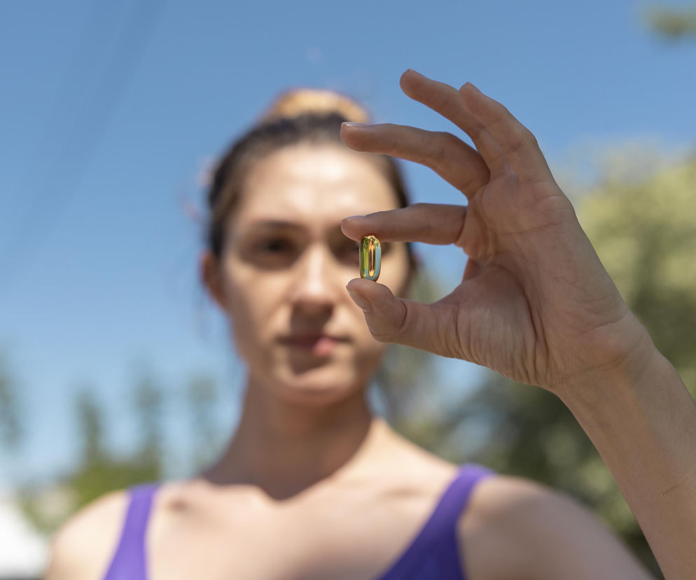 Kobieta trzyma w palcach jedną kapsułkę suplementu witaminy D w otoczeniu zieleni i błękitnego nieba. Dowiedz się, czy witamina D chroni przed cukrzycą typu 2, czytając artykuł na Poradnik Zdrowie.