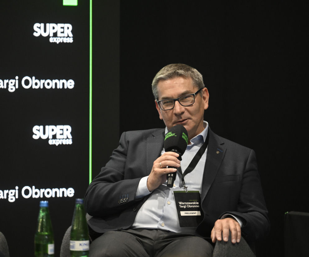 Mężczyzna w okularach i garniturze, z mikrofonem w dłoni, siedzi podczas debaty na Warszawskich Targach Obronnych. Ma na sobie identyfikator „PRELEGENT”. Tło zawiera loga SUPER express i Warszawskie Targi Obronne. Więcej na ten temat przeczytasz na naszym portalu.