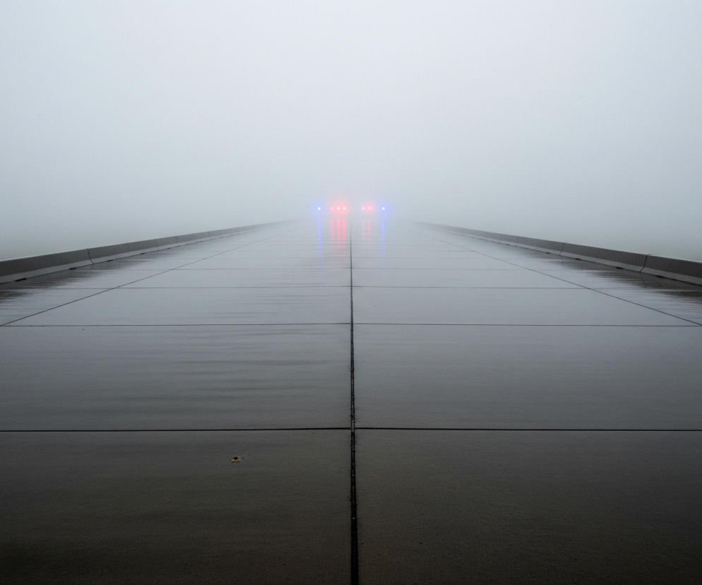 Długa, prosta droga lub pas startowy rozciąga się w głąb obrazu, zanikając w gęstej, szarej mgle. Powierzchnia jest ciemnoszara i błyszcząca, sprawiając wrażenie mokrej i odbijającej światło. W oddali, na horyzoncie, widoczne są cztery jasne światła – dwa czerwone pośrodku i dwa niebieskie po bokach, wszystkie lekko rozmyte przez mgłę. Po obu stronach drogi biegną niskie, ciemne krawężniki, które również znikają w mgle.